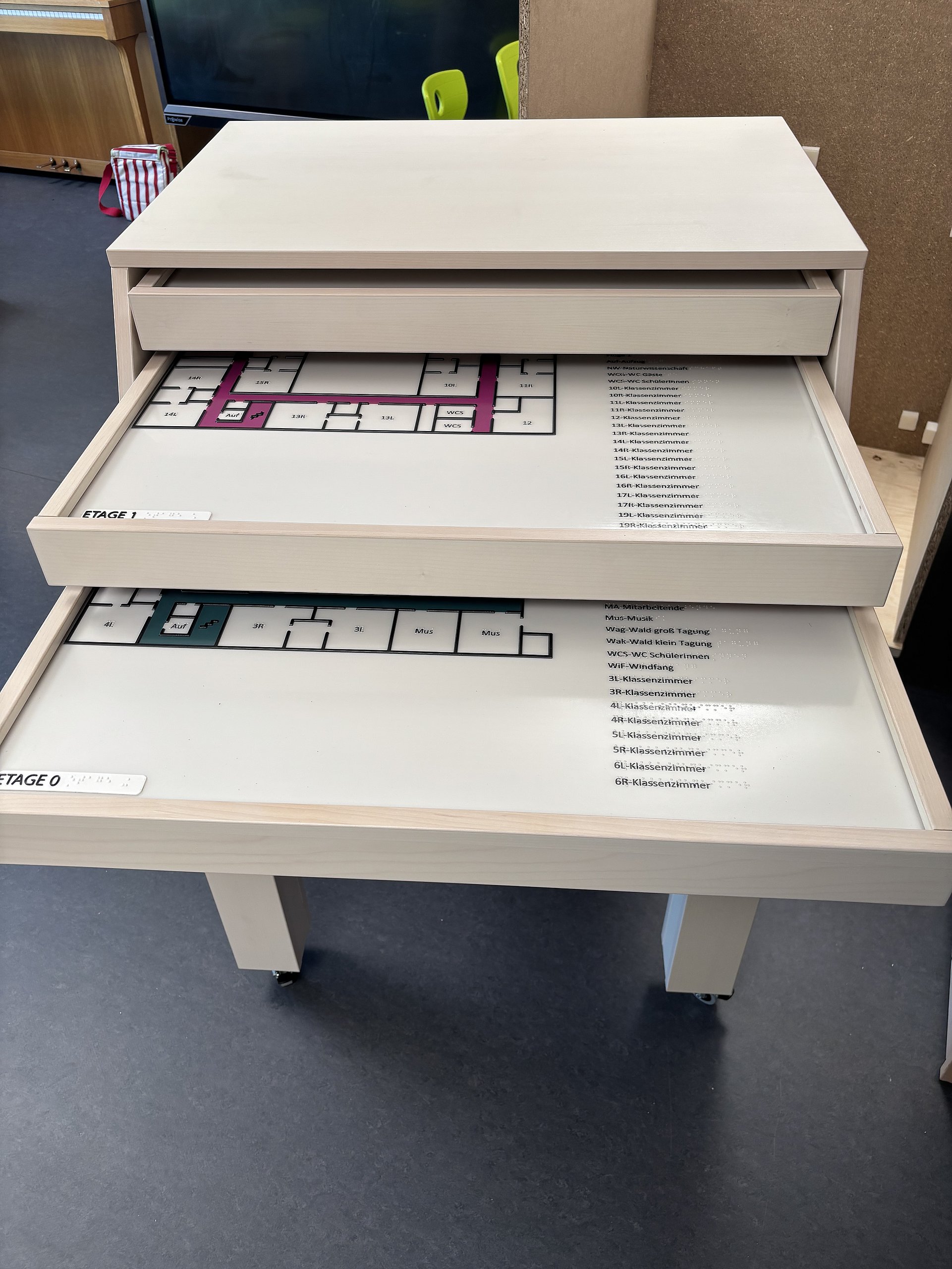 Ein Modellmöbel mit mehreren flachen Schubladen auf Rollen steht in einem Raum. Drei Schubladen sind teilweise herausgezogen, in jeder liegt ein Grundrissplan mit Räumen und Legende. Die Pläne wirken wie Etagenpläne eines Gebäudes, mit rechteckig eingezeichneten Räumen und Nummerierungen.