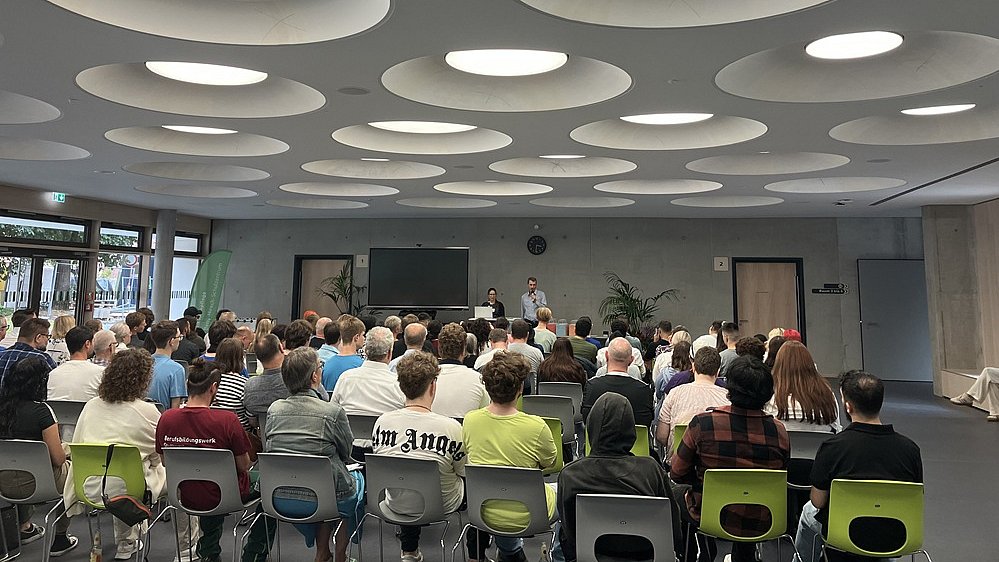 In der Aula des Betty-Hirsch-Schulzentrums sitzen zahlreiche Menschen auf Stühlen in Stuhlreihen. Sie schauen alle nach vorne, wo ein Smartboard aufgebaut ist. Rechts daneben stehen eine Frau und ein Mann mit Mikrofon in der Hand.