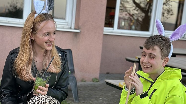 Eine Frau und ein junger Mann sitzen nebeneinander in Stühlen am Lagerfeuer, die tragen Haarreife mit Hasenohren und halten einen Stock mit Stockbrot über das Lagerfeuer. Die Frau hält einen Mocktail in der Hand.