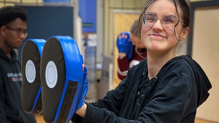 Eine Jugendliche hält vor sich zwei blaue Standboxsäcke in der Hand und lächelt in die Kamera.