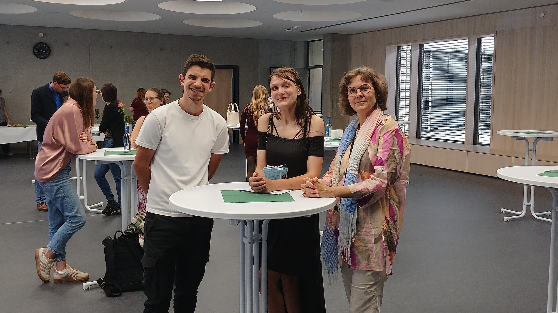 Ein junger Mann in weißem T-Shirt, eine junge Frau in schwarzem Kleid und eine Frau in rosafarbener Bluse stehen nebeneinander an einem weißen Stehtisch und schauen in die kamera.