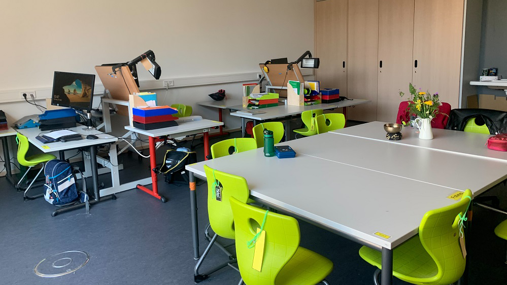 Ein eingerichtetes Klassenzimmer mit weißen Tischen, grünen Stühlen, PCs, Schulrucksäcken.