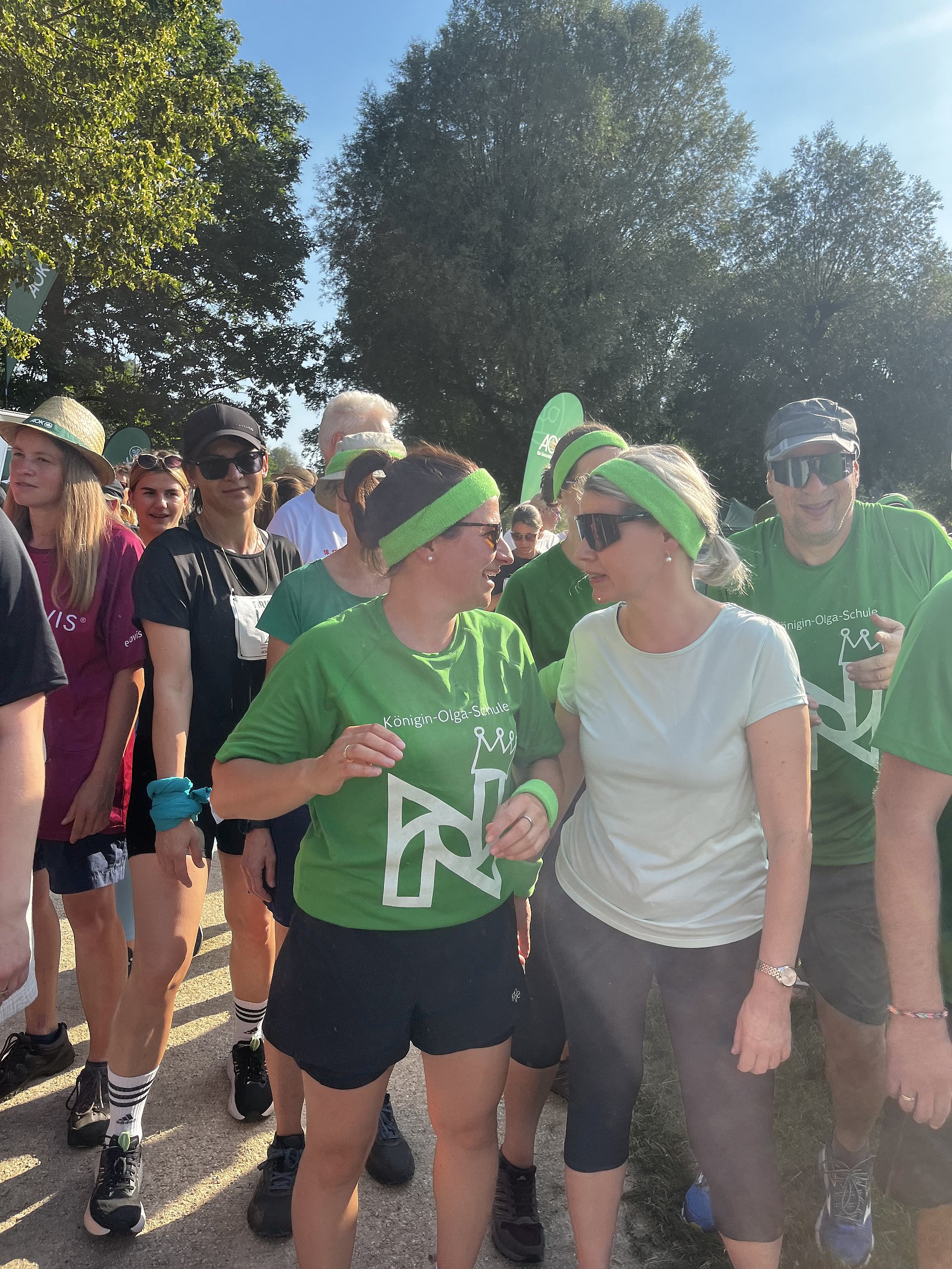 Eine Gruppe von Menschen bei einer Lauf- oder Wanderveranstaltung im Freien. Im Vordergrund stehen zwei Frauen nebeneinander und unterhalten sich lächelnd. Die Frau links trägt ein grünes Stirnband, ein grünes T‑Shirt mit weißer Aufschrift „Königin-Olga-Schule“ und einem stilisierten Logo darauf, dazu dunkle Shorts und Sportschuhe. Die Frau rechts trägt ein helles T‑Shirt, eine dunkle dreiviertellange Sporthose, Sonnenbrille und ein hellgrünes Stirnband, ihre Haare sind zum Zopf gebunden.
