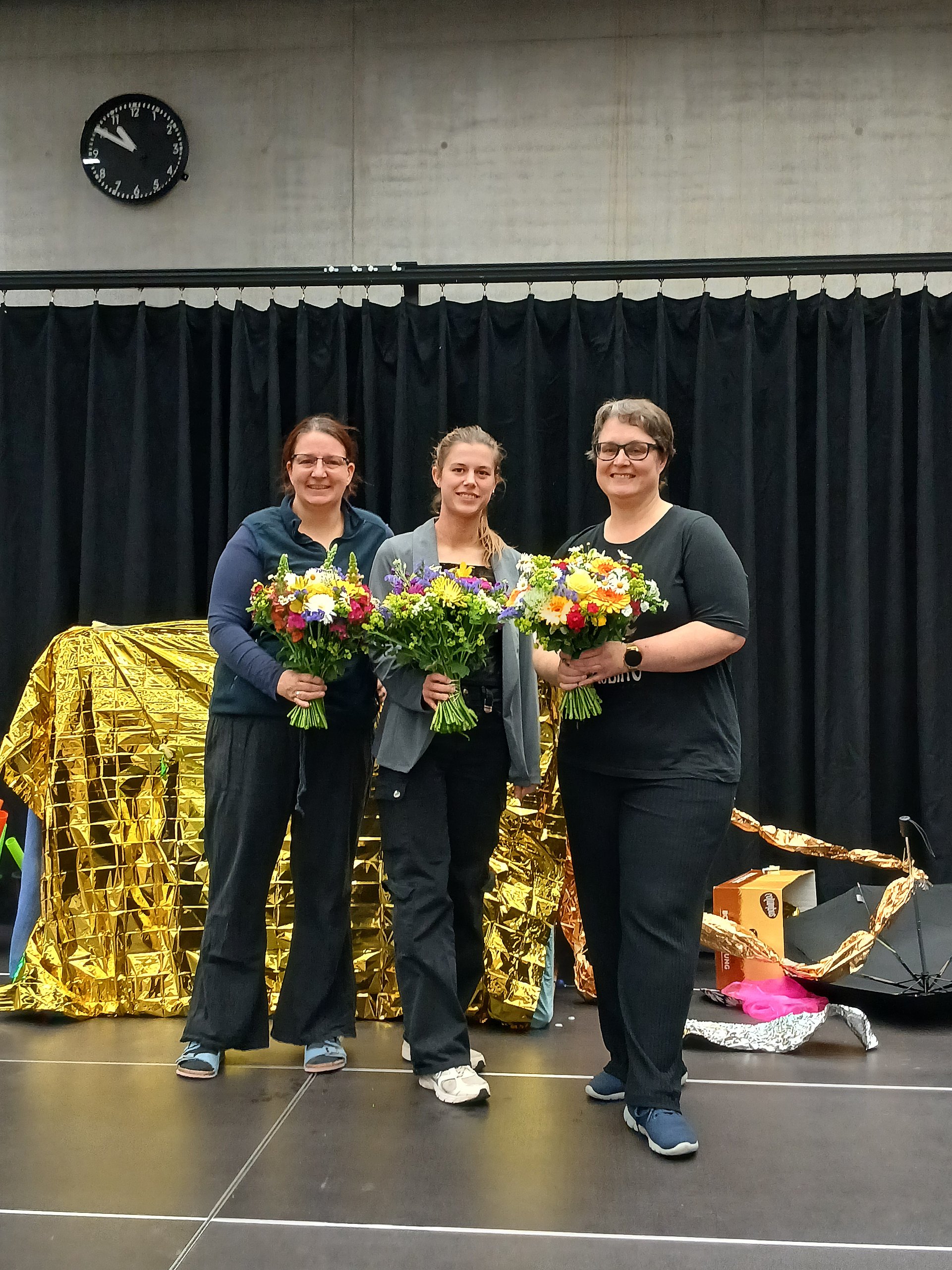Drei Frauen stehen nebeneinander auf einer Bühne und halten bunte Blumensträuße in den Händen. Alle drei lächeln in Richtung Kamera. Die Frau links trägt eine dunkle Hose, ein dunkelblaues Oberteil mit Kapuze und helle Sportschuhe; sie hat hellen Hautton und dunkle, zurückgebundene Haare. Die Frau in der Mitte hat hellen Hautton, mittelblonde, nach hinten gebundene Haare, eine dunkle Hose, ein dunkles Oberteil und darüber eine offene, hellere Jacke sowie helle Sportschuhe. Die Frau rechts ist etwas kräftiger gebaut, hat hellen Hautton, kurze dunkle Haare, eine dunkle Hose, ein dunkles T‑Shirt und dunkelblaue Schuhe; sie trägt eine Armbanduhr am linken Handgelenk. Die Blumensträuße sind groß und gemischt, mit vielen Farben wie Gelb, Weiß, Lila, Rot und Grün. Hinter den Frauen hängt ein schwarzer Vorhang, davor liegt links eine große, goldglänzende Decke oder Folie, die gefaltet und drapiert ist.