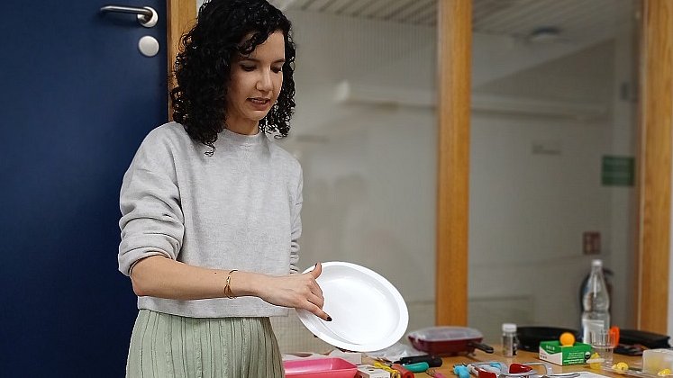 Eine Frau mit schwarzen Locken, weißer Bluse und türkisfarbenem Rock steht im Raum vor einer blauen Tür. Sie hält einen weißen Teller in der Hand.