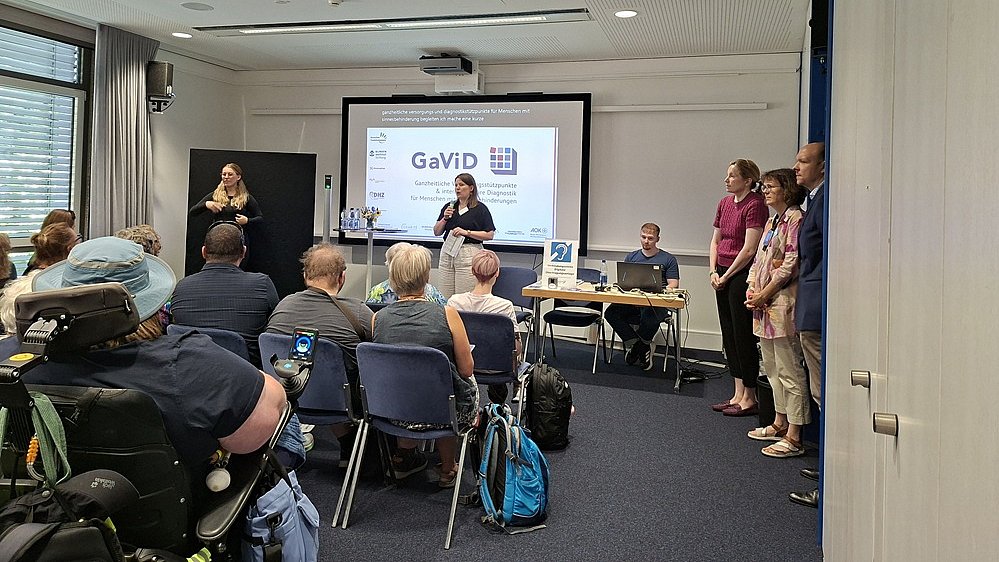 Konferenzraum mit sitzendem Publikum und einer Präsentation vorne. Mehrere Reihen mit Menschen sind von hinten zu erkennen, einige sitzen auf Stühlen, eine Person sitzt in einem elektrischen Rollstuhl, daneben stehen Rucksäcke und Taschen. Vorne im Raum steht eine Projektionsfläche mit einer Präsentationsfolie; auf der Leinwand ist groß das Wort „GAVID“ zu lesen sowie mehrere Logos und kleinere Texte. Vor der Leinwand befindet sich ein Tisch mit Unterlagen, dahinter sitzt eine Person. Rechts vor dem Tisch stehen mehrere Personen, teils einander zugewandt, als würden sie sprechen oder etwas vorstellen.  