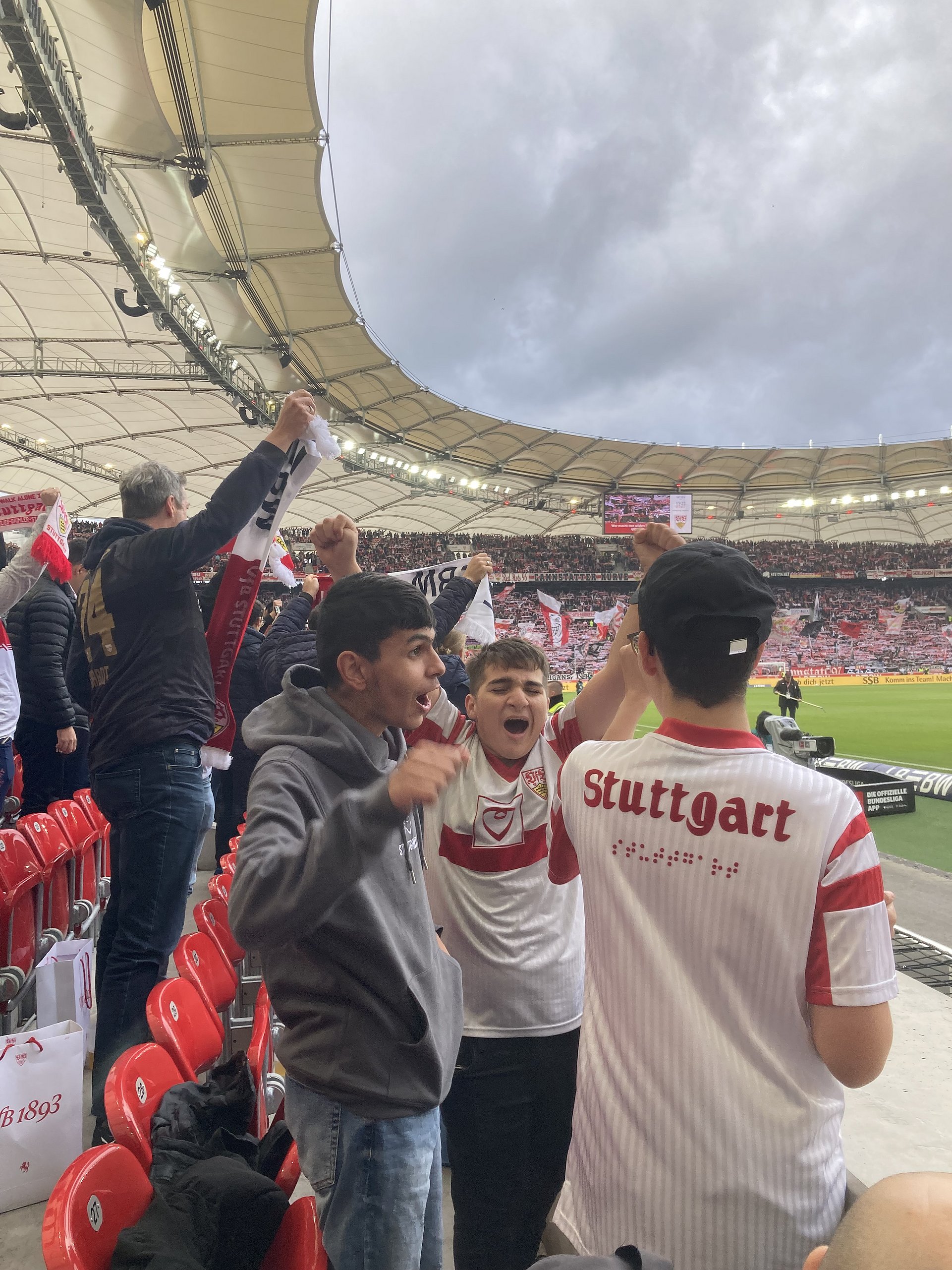 Fußballfans im Stadion: Das Bild zeigt mehrere junge Männer, die im Stadion auf roten Sitzen jubeln. Zwei von ihnen tragen Trikots des VfB Stuttgart – auf dem Rücken eines Trikots steht groß „Stuttgart“. Im Hintergrund sieht man weitere Zuschauer, das Stadiondach und den Rasen.