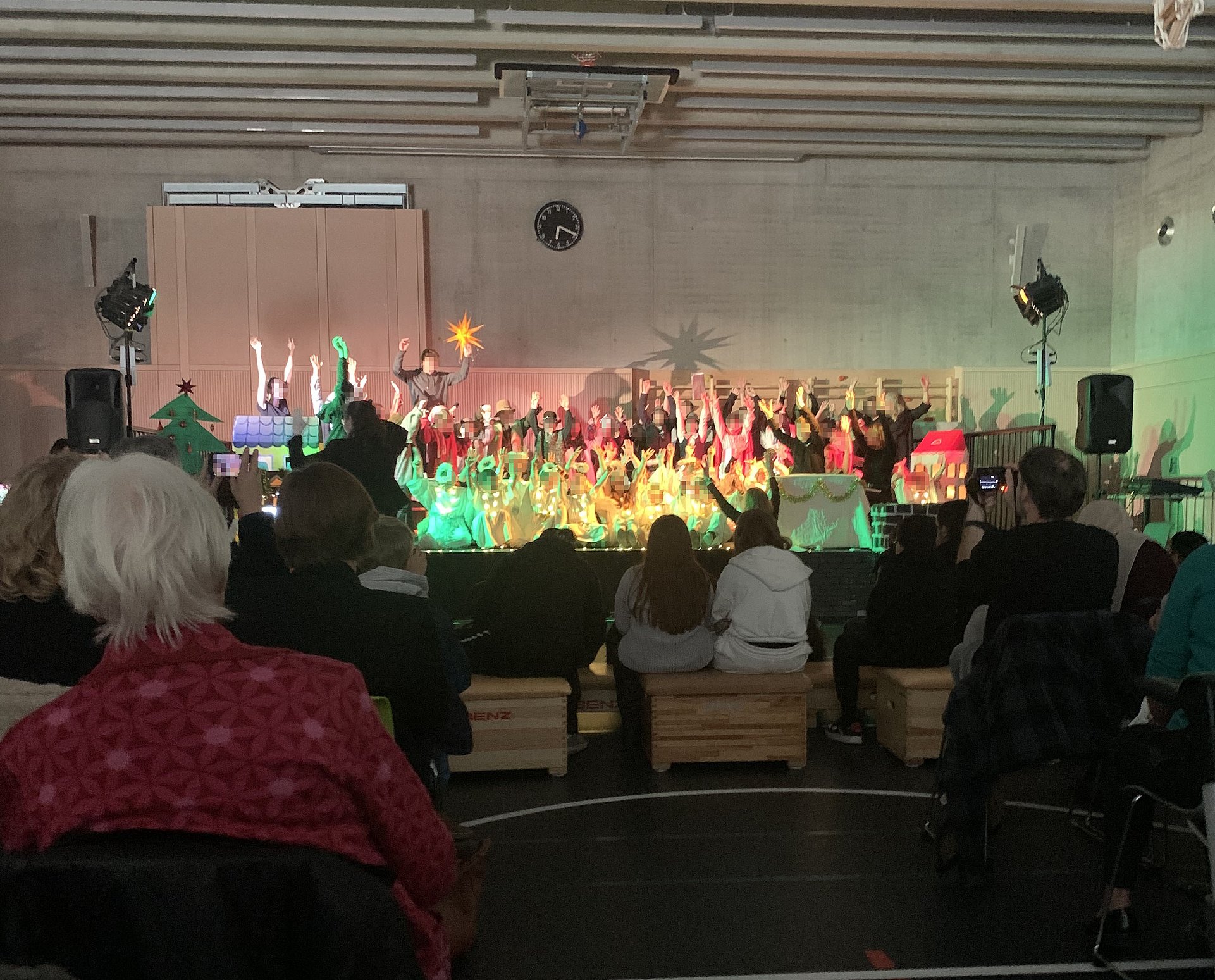 In der abgedunkelten Sporthalle des Schulzentrums sitzen viele Zusachauer vor einer Bühne die hell und bunt beleuchtet ist. Auf der Bühne stehen Schülerinnen und Schüler als Engel verkleidet