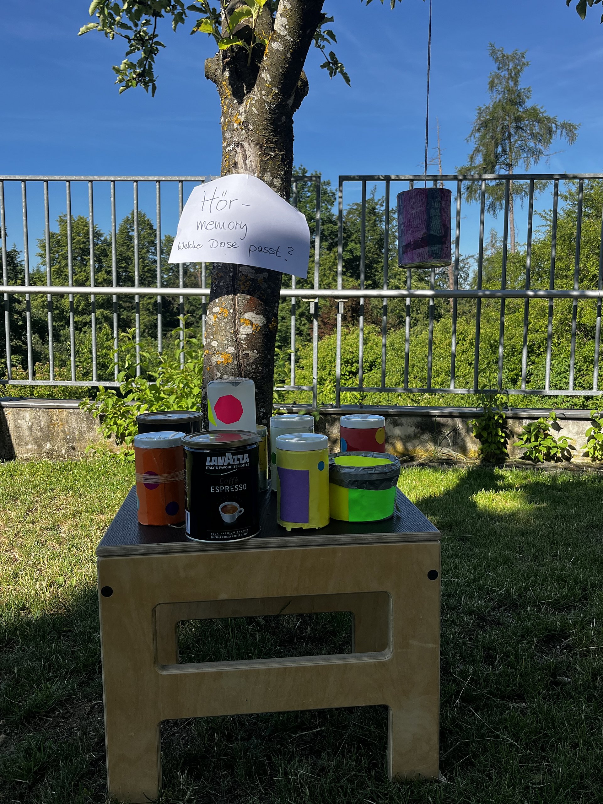 Ein kleiner Holzhocker mit mehreren bunt beklebten Dosen darauf im Freien. Auf dem Hocker stehen von links nach rechts: Hinter dem Hocker steht ein Baum. An dem Baum ist ein weißes Blatt Papier befestigt, darauf steht handgeschrieben:„Hör – Memory Welche Dose passt?“ Rechts vom Baum hängt an einer Schnur eine lila Dose oder ein lila beklebter Zylinder in ungefähr Augenhöhe, offenbar als Teil des Spiels.