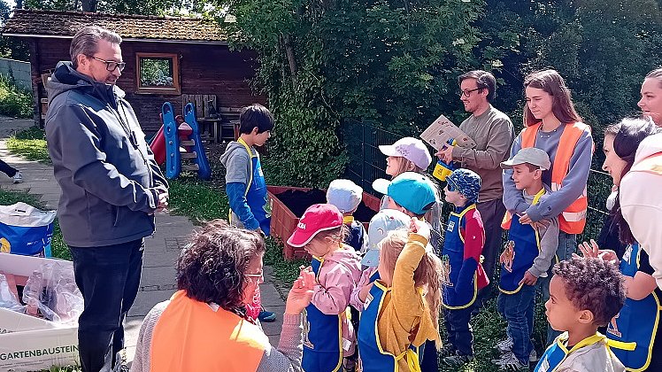 Eine Gruppe Kinder und mehrere Erwachsene stehen in einem Garten, die Kinder tragen gelb-blaue Schürzen.