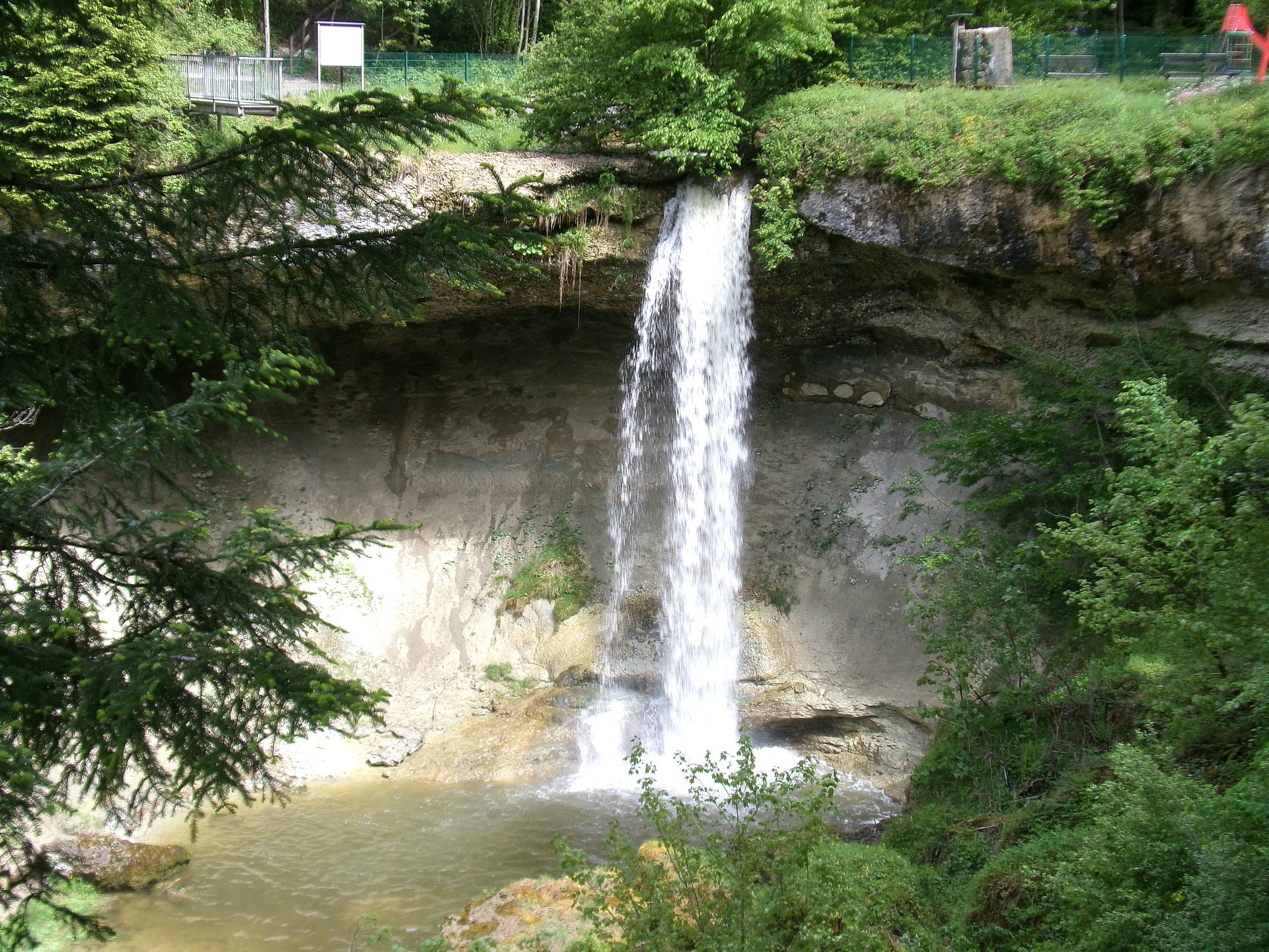 Ein Wasserfall, der von einer Felskante in ein darunterliegendes Becken stürzt. Das Wasser fällt in einem breiten, weißen Schleier die graue Felswand hinunter