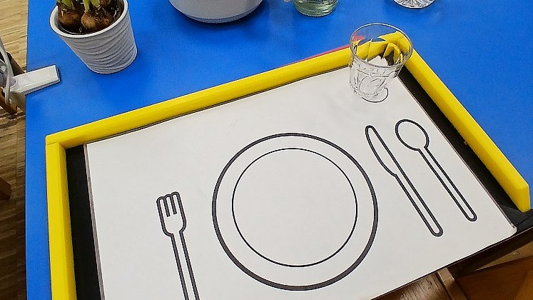 Auf einem blauen Tisch steht ein schwarzes Tablett mit gelbem Rand, darauf eine weiße Mappe, auf der mit schwarz die Umrisse eines Tellers und Besteckt gemalt sind. Rechts oben steht ein Glas.