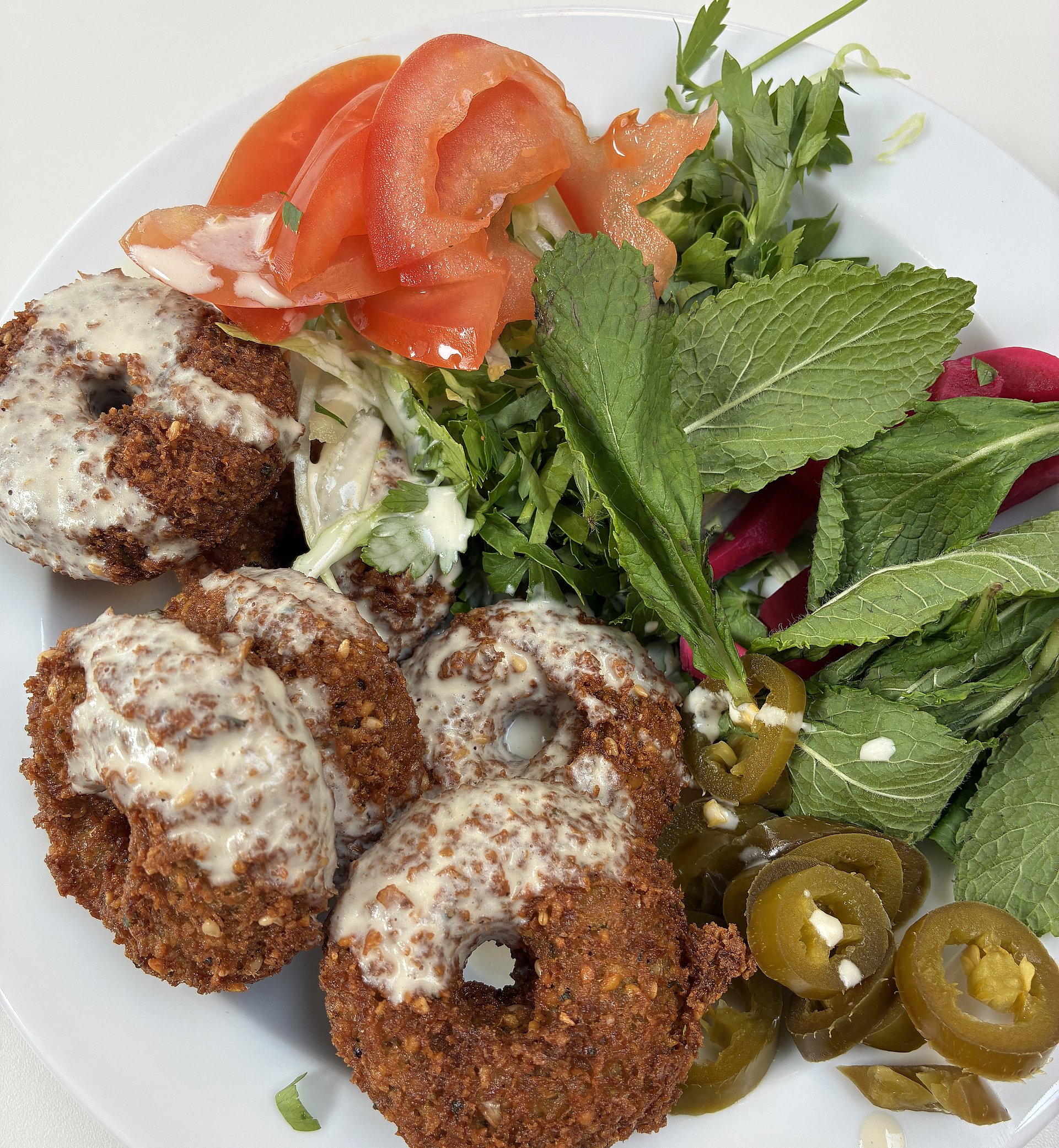 Auf einem Teller sind Falaffelbällchen mit Salat, Tomaten und Oliven. Darüber weiße Soße