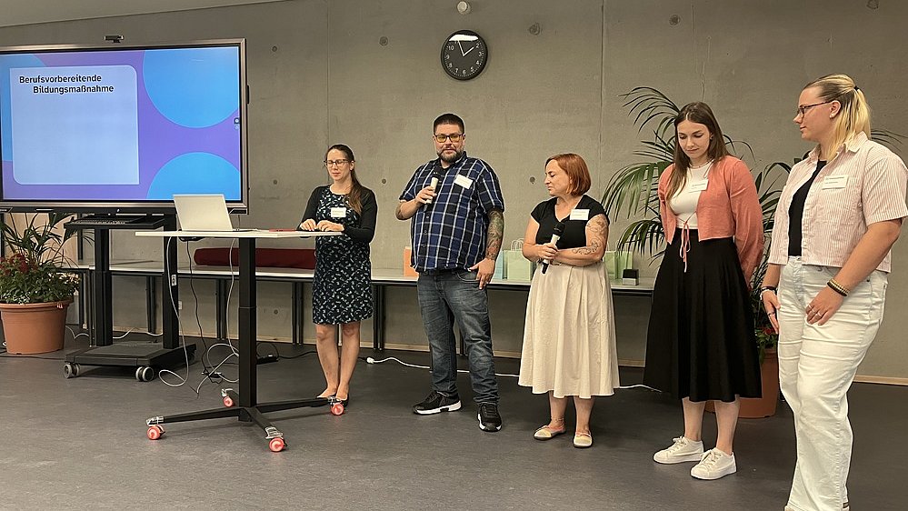 Eine Frau mit schwarzem Kleid und schwarzer Weste steht an einem Stehtisch neben einem Smartboard mit einer Präsentation darauf. Neben ihr steht ein Mann in Jeans und Hemd und einem Mikrofon in der Hand, rechts daneben eine Frau mit beigem Rock, schwarzer Bluse und Mikrofon in der Hand. Daneben eine Frau in schwarzem Rock und rosafarbener Bluse sowie eine Frau in weißer Hose und weißer Bluse.