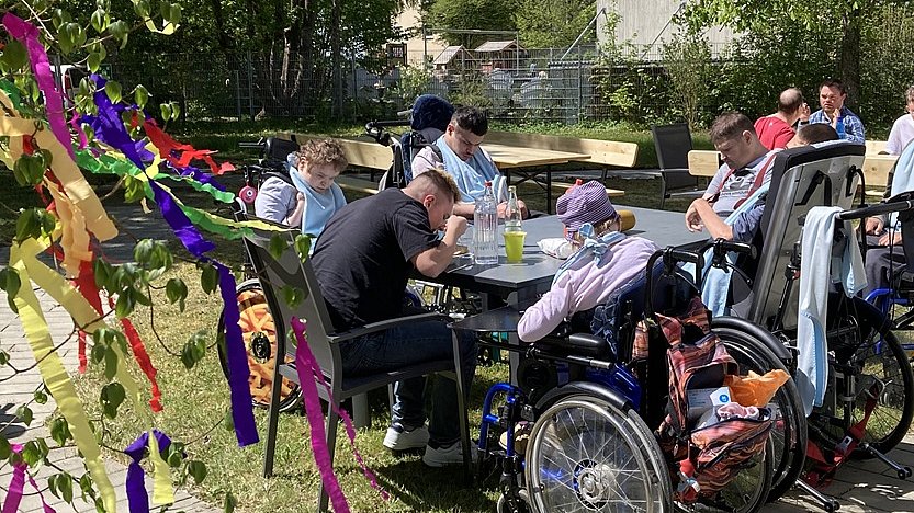 Auf einer Wiese stehen Tische und Stühle, an denen Menschen sitzen und essen. Einige davon sitzen im Rollstuhl. Im Hintergrund sind Bäume und Büsche zu sehen, links wehen bunte Papierfahnen ins Bild hinein.