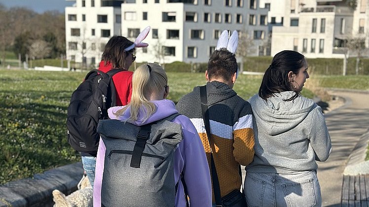 Vier Jugendliche und ein Hund laufen nebeneinander über einen Weg, im Bild hinten sind große weiße Häuserblocks, links und rechts grüne Wiese.