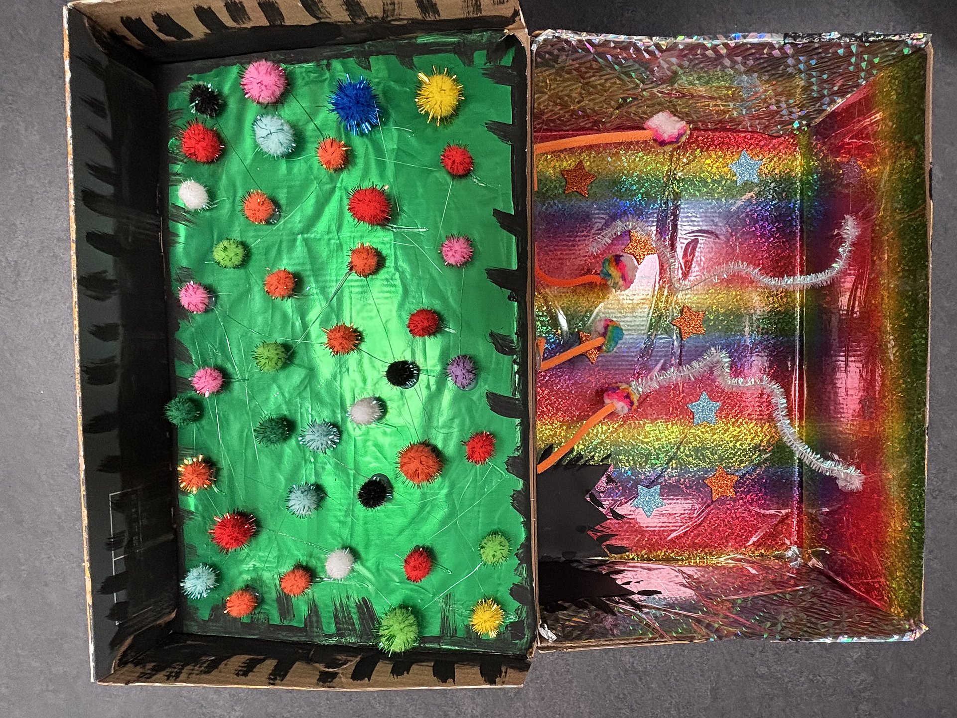 Ein aufgeklappter bunt gestalteter Pappkarton. Die linke Seite ist grün, die reche in Regenbogenfarben