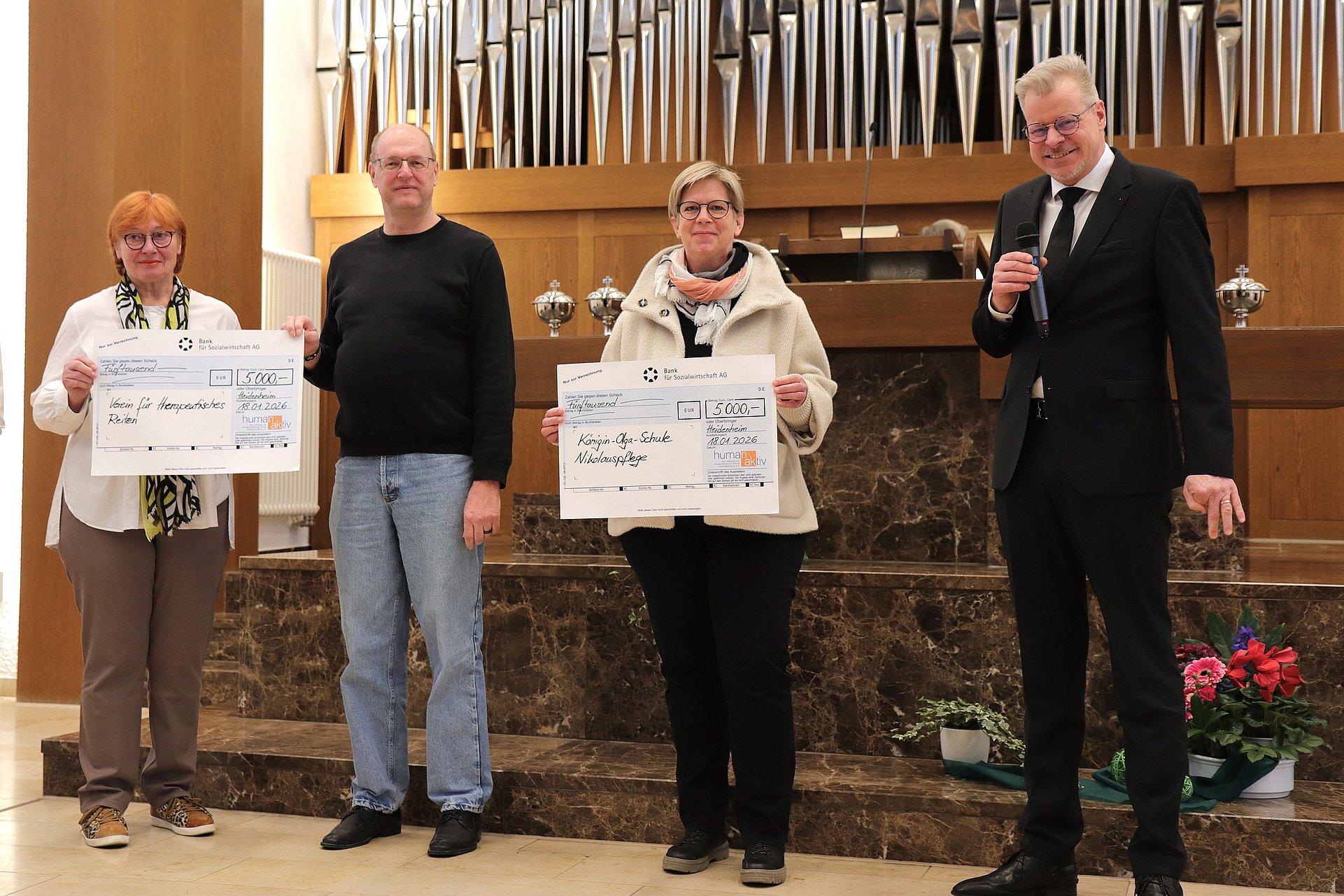 Zwei Frauen und zwei Männer stehen vor einem Altar in der Neuapostolischen Kirche. Hinter ihnen ist eine große Orgel zu sehen. Die Frauen  halten einen großen Scheck in den Händen