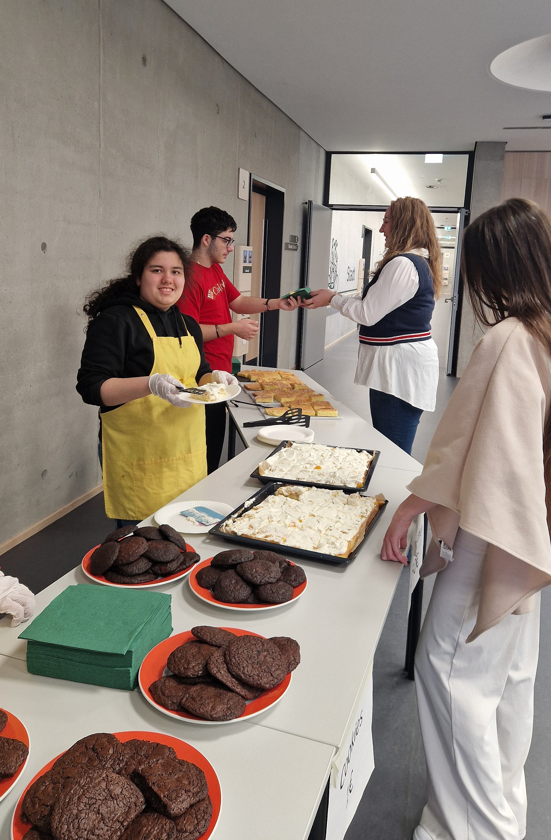 Eine Schülerin und ein Schüler stehen in einem Flur hinter aneinander gereihten Tischen. Darauf stehen viele verschiedene Kuchen. Der Schüler reicht einer Lehrerin ein Stück Kuchen. Vor den Tischen steht eine Schülerin.