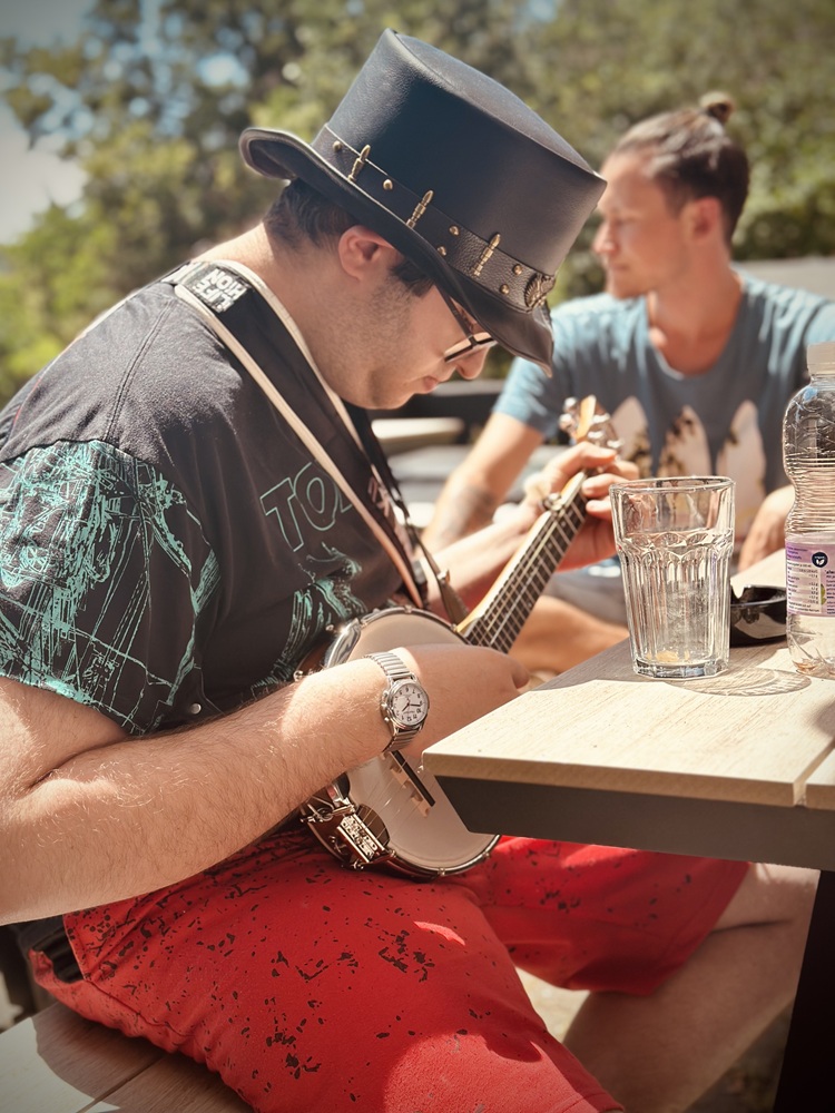 Ein junger Mann der eine Brille und einen Zylinder trägt, sitzt an einem Tisch im Freien und spielt auf der Gitarre. Auf dem Tisch stehen Gläser und Flaschen. Im Hintergrund sitzt noch ein Mann