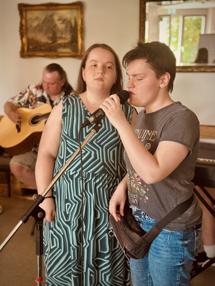 Zwei junge Frauen stehen nebeneinander. Die rechte Frau singt in ein stehendes Mikrofon. Im Hintergrund sitzt ein Mann und spielt die Gitarre