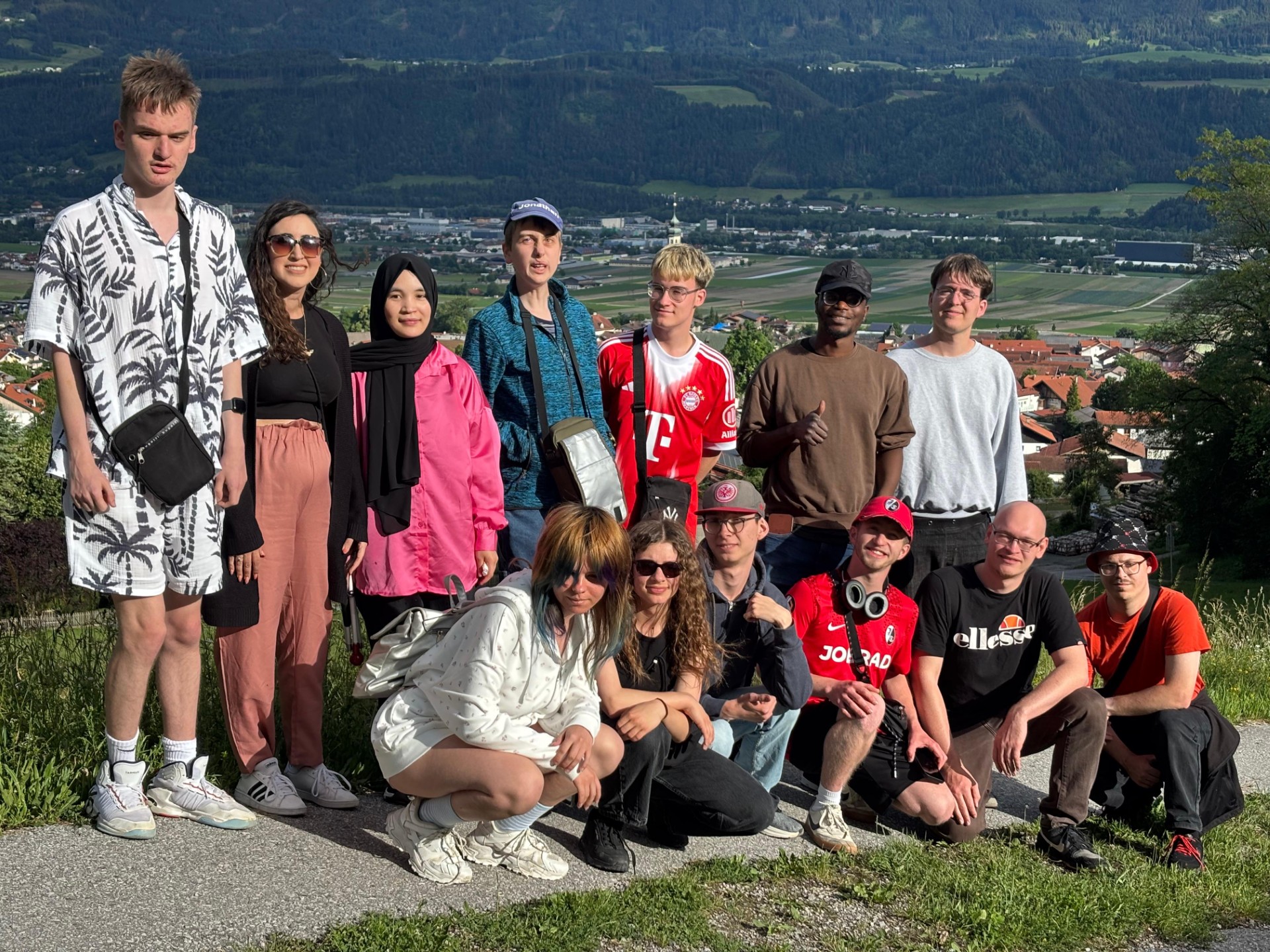 Eine größere Gruppe junger Menschen sitzt und steht im Freien vor einer Berglandschaft. Im Vordergrund sitzt eine Reihe von Personen auf einem niedrigen Mäuerchen oder Steinrand. Alle wirken entspannt und posieren nah beieinander für ein Gruppenfoto. Im Hintergrund erstreckt sich ein weitläufiges Tal mit Feldern, einem Ort mit orangefarbenen Dächern und hohen grünen Bergen dahinter.