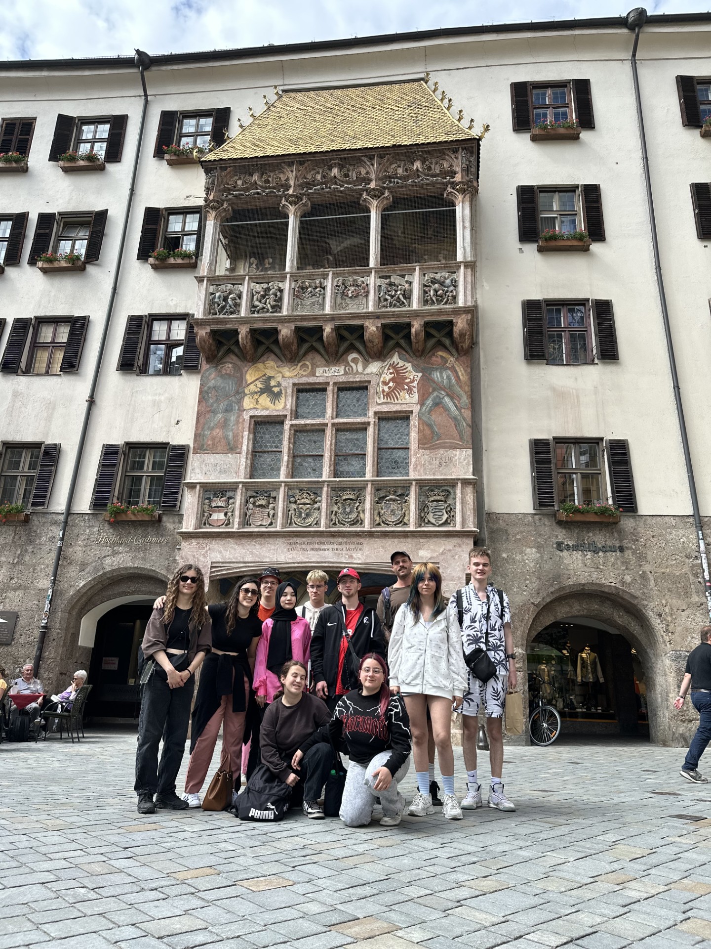 Eine Gruppe junger Menschen vor einer historischen Hausfassade mit reich verziertem Erker. Vor dem Gebäude steht eine größere Gruppe, etwa zwölf Personen, die in zwei Reihen posieren: einige hocken vorne auf dem Pflaster, die anderen stehen dahinter. Die Kleidung ist modern und lässig – überwiegend dunkle Töne, dazu einzelne auffällige Farben wie ein leuchtend pinkes Outfit und eine weiße, langärmelige Kapuzenjacke mit kurzen Hosen. Mehrere Personen tragen Turnschuhe, einige haben Rucksäcke oder Umhängetaschen. Die Stimmung wirkt freundlich und wie bei einem Ausflugsfoto.